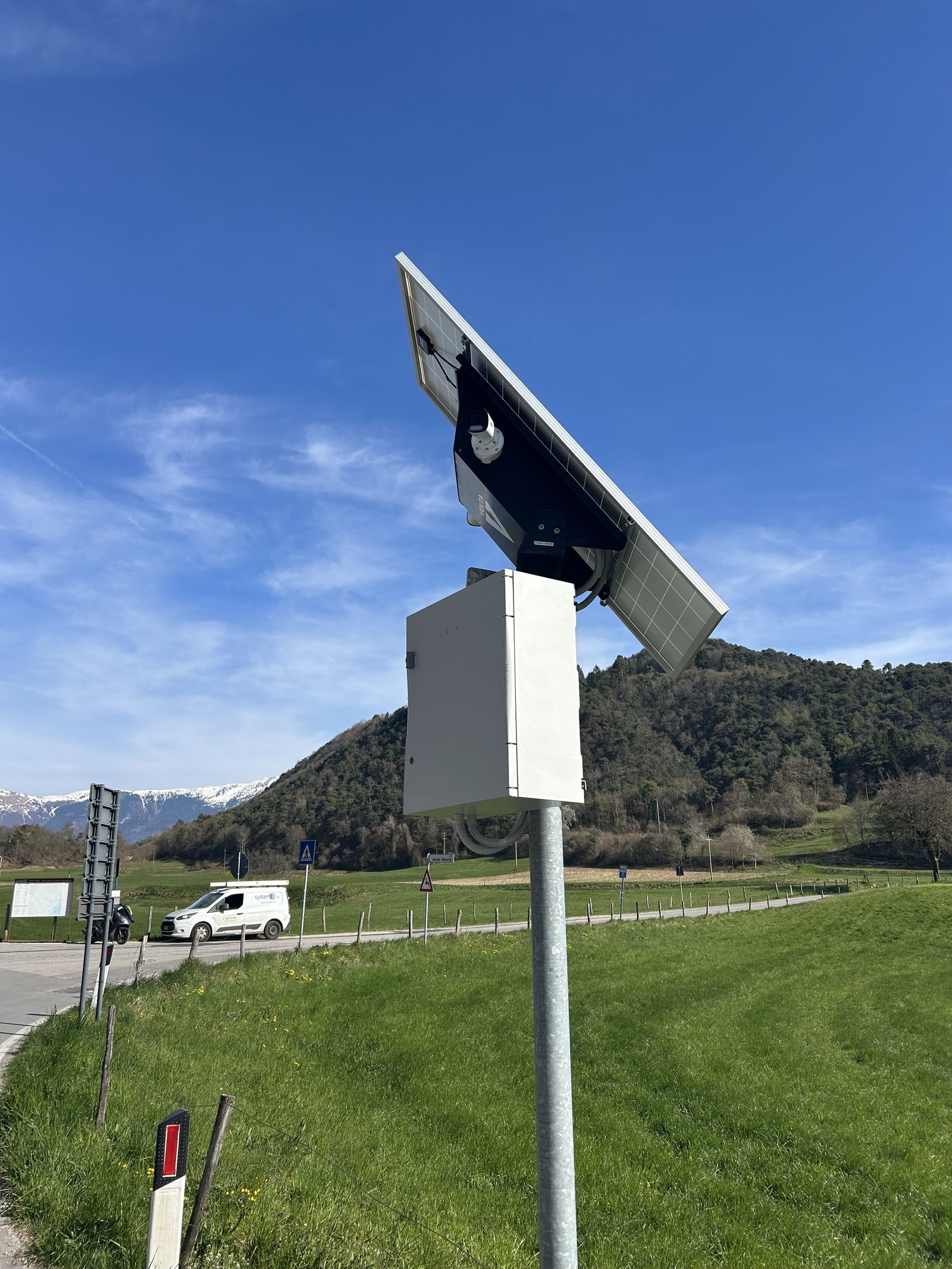 Postazione di videosorveglianza off-grid a Tremosine sul Garda: pannello solare, cassa batterie e telecamera montati su palo, con sullo sfondo il paesaggio prealpino e le vette innevate. Il furgone di servizio System-i sulla strada